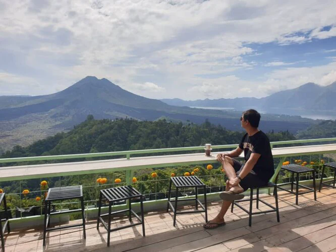 A visitor with a cup of coffee at a Kintamani viewpoint looking across to Mount Batur