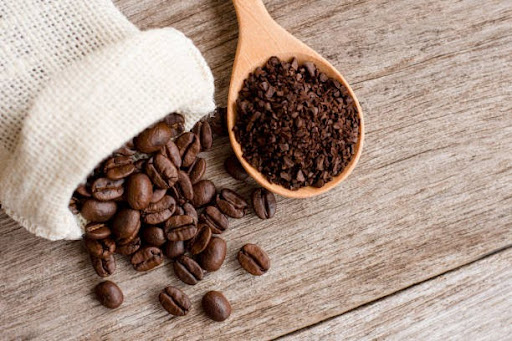 Roasted coffee beans spilling from a jute sack beside ground coffee on a wooden spoon